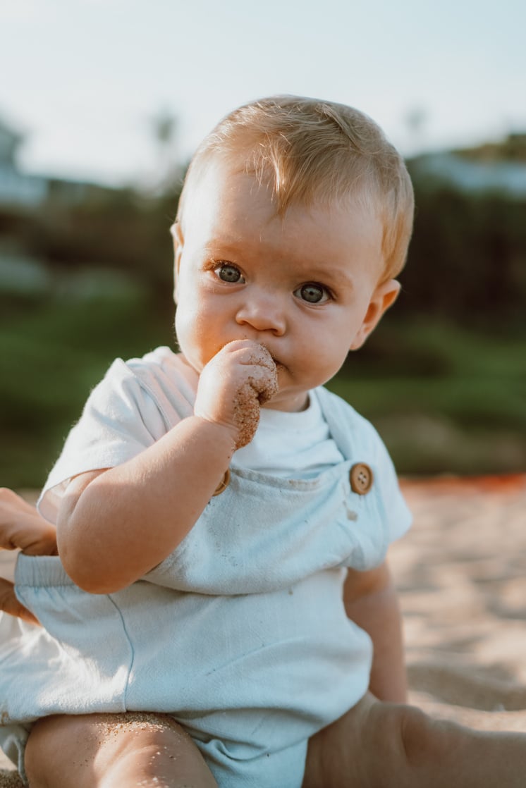 a baby sitting on the beach with his hand in his mouth